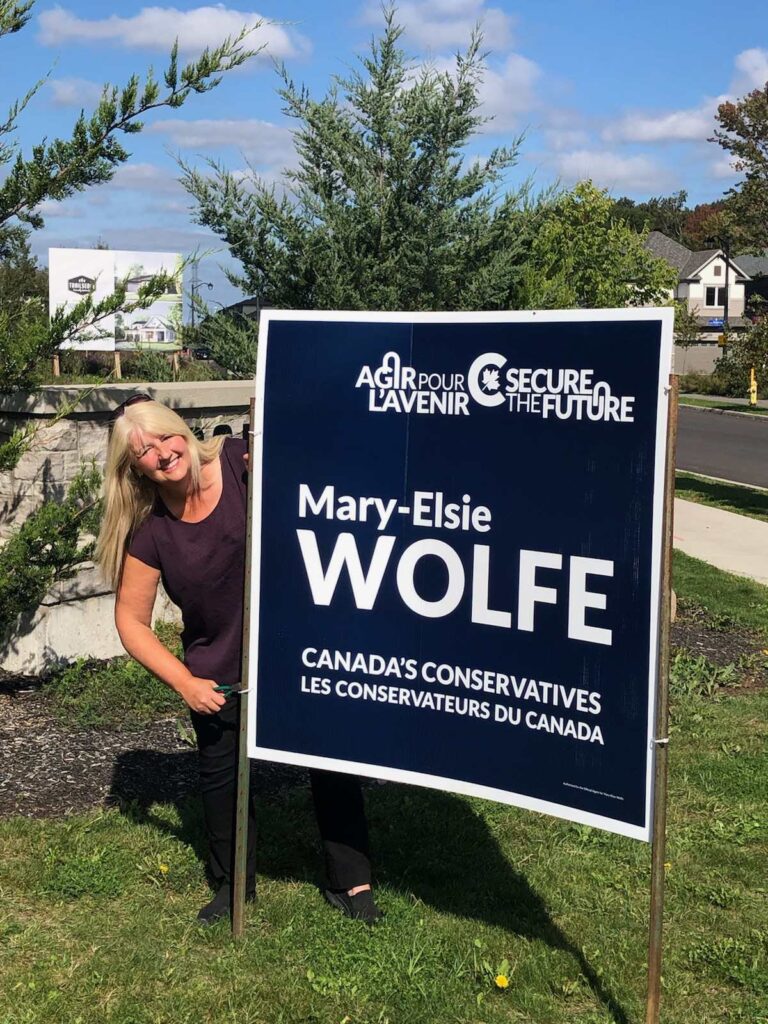 Mary-Elsie Wolfe stands behind a large campaign sign. 